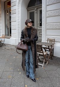 Casaco longo com padrão de leopardo e mangas de couro, suéter preto, jeans azuis de perna larga, bolsa castanha e protetores de orelhas, em pé numa calçada da cidade.