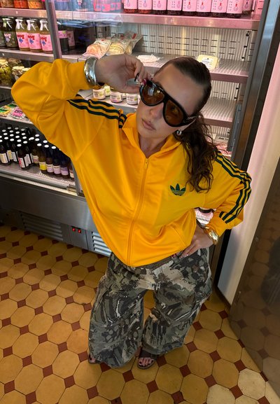Chaqueta de chándal amarilla con cremallera y rayas verdes, combinada con pantalones cargo anchos de camuflaje, frente a un colorido fondo de tienda.