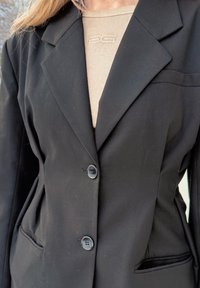 Γυναίκα που φοράει μαύρο σακάκι με δύο κουμπιά κουμπωμένα πάνω από μια μπεζ ριμπ παλτό κεντημένο με διακριτικό λογότυπο στο ντεκολτέ.