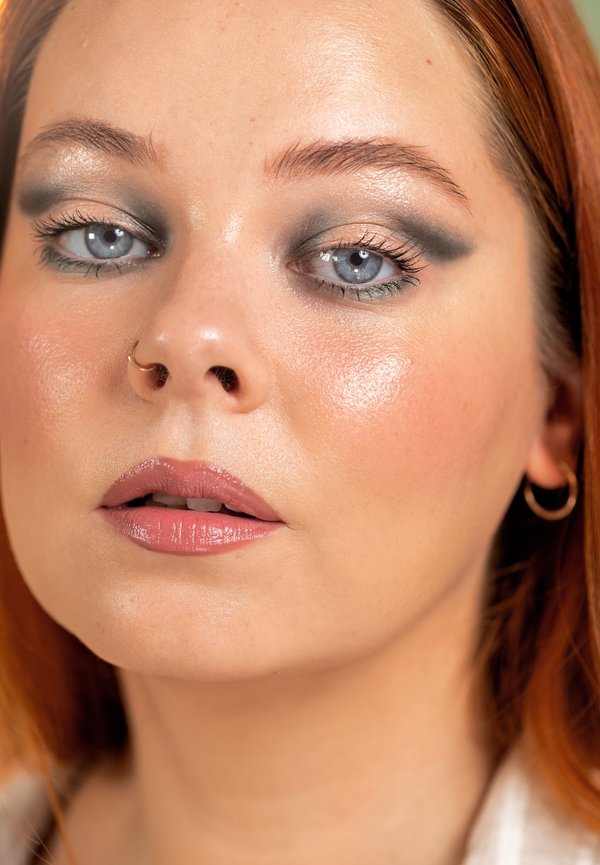 Nahaufnahme einer Frau mit heller Haut, roten Haaren, blauen Augen, Smoky-Eye-Make-up, glänzenden rosa Lippen, Nasenring und goldenen Creolen.