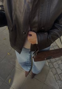 Črna usnjena jakna s pritrdilnimi gumbi, lahka jopica pod njo, modri široki kavbojke, rjava semiš torba in špičasti rjavi škornji.