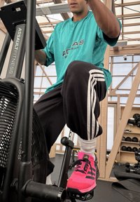 Rower treningowy Assault Fitness z matową czarną ramą. Użytkownik ma na sobie turkusową koszulkę i czarne dresy z białymi paskami oraz jaskraworóżowe trampki.