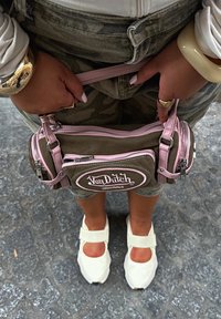 Person der holder en brun og lyserød Von Dutch taske, iført camouflagebukser, hvide sko, guld- og kremfarvede armbånd, stående på brosten.