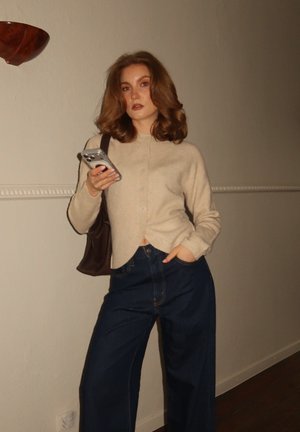 Joven con cabello castaño hasta los hombros está de pie en interiores sosteniendo un teléfono inteligente, vistiendo un suéter beige, pantalones anchos oscuros y un bolso de hombro.