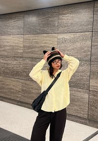 Chemise jaune surdimensionnée à boutons, sac noir, pantalon noir, lunettes rondes, portant un bonnet en tricot rayé avec des oreilles de chat. Intérieur avec des murs en pierre.