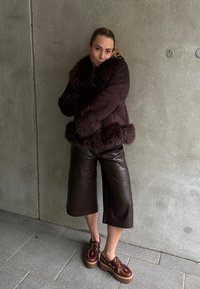 Veste en fausse fourrure marron avec un col épais, associée à des culottes en cuir marron et des chaussures à plateforme bordeaux avec lacets.