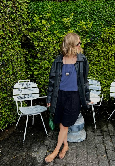 Mujer con gafas de sol, chaqueta de cuero negra, top azul marino y falda negra de pie junto a sillas blancas de metal con una pared cubierta de vegetación detrás.