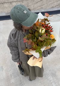 Grey bomber jacket, olive skirt, green cap with logo, holding a mixed flower bouquet with orange and yellow accents and foliage.