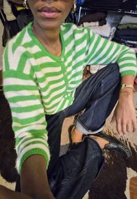 Žaliai ir baltai dryžuotas megztinis su sagomis, derinamas su tamsiai mėlynais džinsais. Juodi sliponai užbaigia aprangą. Fone matyti sulankstyti drabužiai.