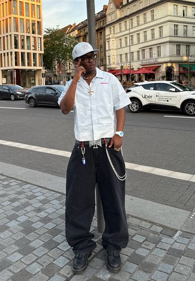 Camisa blanca de mangas cortas con letras rojas, pantalones negros holgados, acentos de cadena, zapatos negros con flecos y una gorra blanca. Entorno urbano.