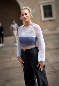 Biała siateczkowa bluzka z długim rękawem, krótka, nałożona na jasnoniebieską dopasowaną bluzkę, w połączeniu z czarnymi, szerokimi spodniami z wysokim stanem. Ręce na biodrach.