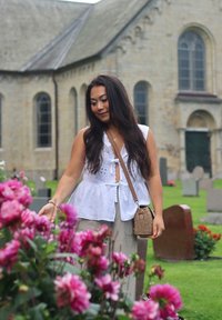 Valge ilma varrukateta pluus dekoratiivsete lõigetega, beežid püksid ja punutud ripp-kott; ümbritsetud roosadest lilledest kalmistu keskkonnas.