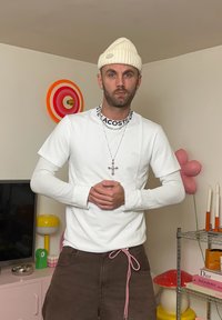 T-shirt branca de manga curta com uma camisa de manga longa canelada por baixo, calças castanhas com um cordão rosa, e um gorro de malha branco. Acessórios de joalharia em prata.