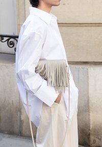 Camicia oversize bianca, pantaloni beige larghi, con una borsa di pelle grigia chiara con dettagli in frangia e tracolla.