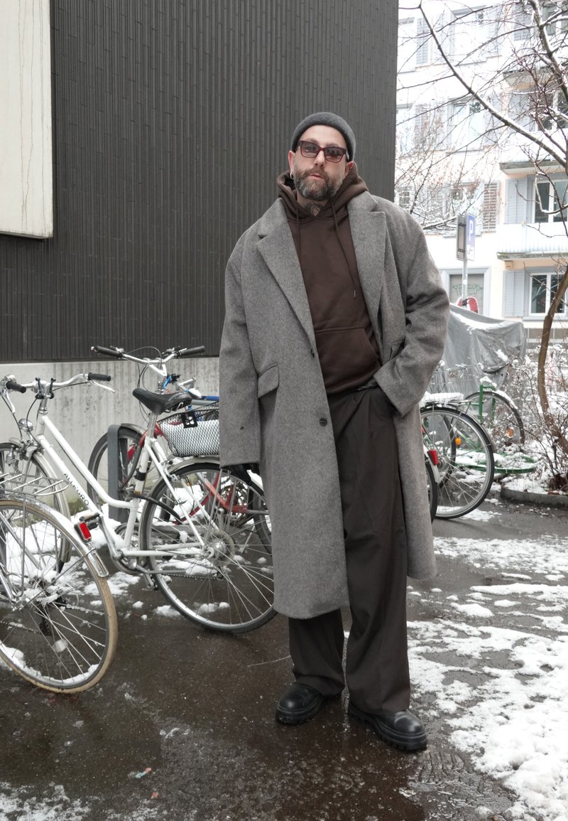 Homme portant un pardessus gris, un sweat à capuche marron, un bonnet et des lunettes de soleil, debout sur un trottoir enneigé et mouillé à côté de vélos stationnés près d'un bâtiment.