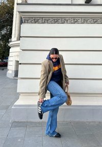 Μπεζ καρό oversized σακάκι, μπλε hoodie, φαρδιές μπλε τζιν, μαύρα sneakers με άσπρες ρίγες, κρατώντας μια μικρή καφέ τσάντα, στέκεται μπροστά σε έναν τοίχο.