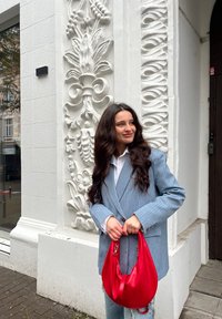 Ανοιχτό μπλε σακάκι με ρίγες πάνω από ένα λευκό πουκάμισο, κρατώντας μια κόκκινη δερμάτινη τσάντα. Το φόντο περιλαμβάνει περίτεχνες λευκές τοιχογραφίες.