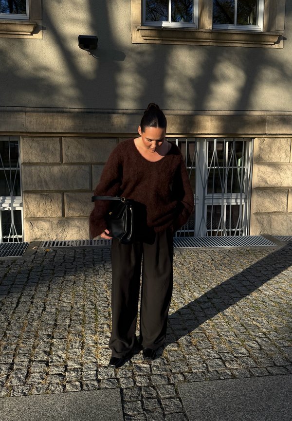 Una mujer con un jersey oscuro y pantalones de pernera ancha mira hacia abajo un bolso negro sobre un pavimento adoquinado, frente a un edificio de piedra.