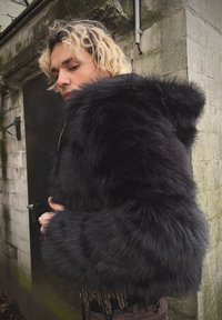 Μαύρο παλτό με κουκούλα, με πλούσια υφή, φαρδιά μανίκια και κλείσιμο με φερμουάρ. Ο σχεδιασμός είναι εφαρμοστός και κατασκευασμένο από συνθετικά υλικά.