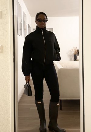 Mujer con chaqueta negra con cremallera, pantalones negros, botas negras altas, gafas de sol y sosteniendo un pequeño bolso negro, de pie en el interior.