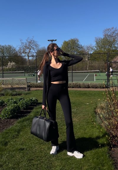 Conjunto atlético negro que incluye un top corto y mallas acampanadas, combinado con un gran bolso de piel negro y zapatillas blancas. Fondo de hierba verde.