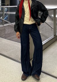 Veste en cuir noire, pull crème, écharpe rouge, jean large bleu, chaussures marron. Détail de couture remarquable sur le jean, debout à l'intérieur.