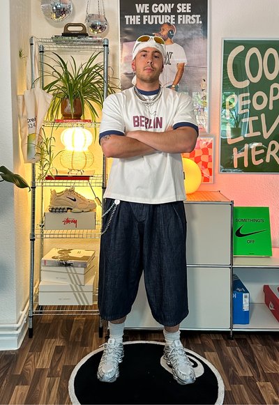 Camiseta blanca con "BERLÍN" en rojo, acentos en azul marino, pantalones cortos de mezclilla oscura, zapatillas plateadas, cadenas en capas y accesorios. Entorno interior con plantas.