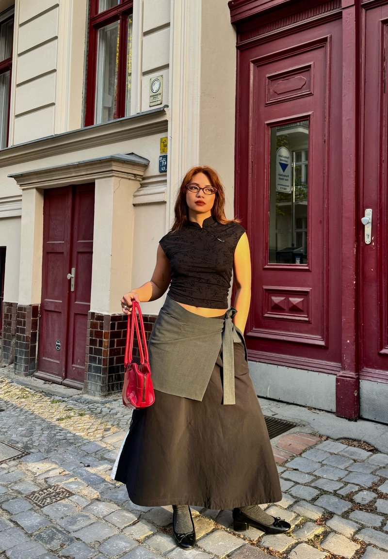 Jeune femme portant un haut noir et une longue jupe portefeuille se tient dans une rue pavée, tenant un sac à main rouge près d'une porte rouge.