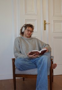 Personne assise sur une chaise en bois avec un rembourrage gris, portant un pull gris clair et un jean bleu, lisant un livre avec des écouteurs.