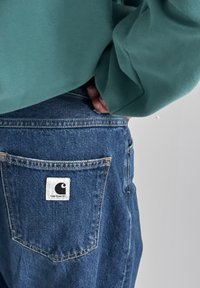 Jean en denim d'un bleu moyen avec une poche arrière arborant une étiquette carrée blanche affichant le logo Carhartt. Sweat-shirt vert clair visible.