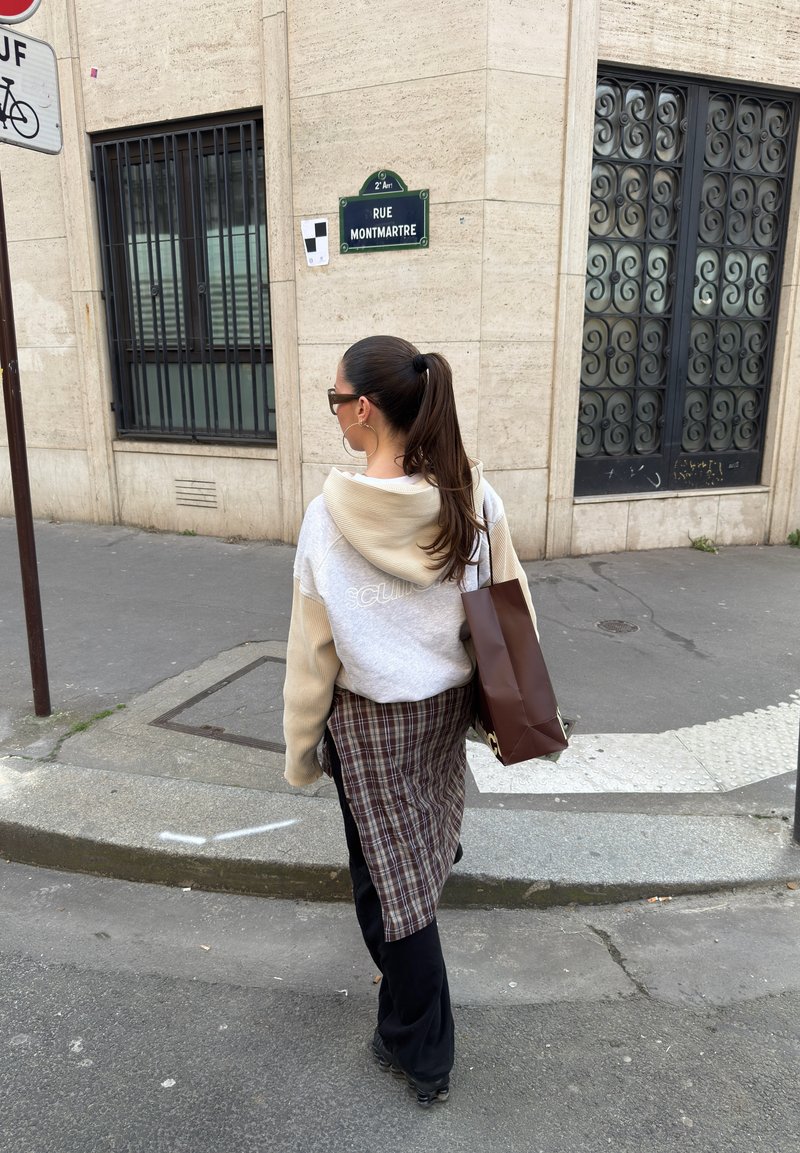 Woman with ponytail wearing sunglasses, beige hoodie, plaid skirt, and black pants holding brown shopping bag walking near Rue Montmartre sign.