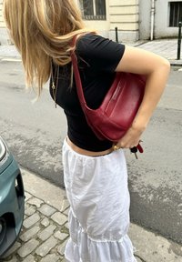 Κόκκινη δερμάτινη τσάντα ώμου με λεία υφή και μινιμαλιστικό σχέδιο. Φορεμένη στον ώμο, συνδυασμένη με μαύρη μπλούζα και λευκή φούστα.