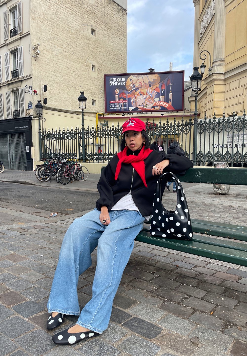 Jeune personne portant une casquette rouge et une écharpe, une veste noire, un jean large, des chaussures et un sac à pois, assise sur un banc vert dans une rue de la ville.