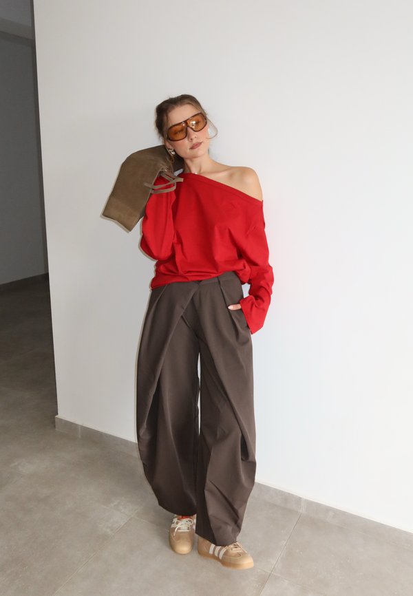 Mujer con top rojo oversize sin hombros, pantalones anchos marrones, zapatillas beige y gafas de sol naranjas, sosteniendo un bolso marrón en el hombro.
