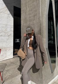 Ένα καφέ σακάκι πάνω από ένα λευκό πουκάμισο, συνδυασμένο με ένα καφέ crop top και πλεκτό γκρι κολάν. Τα αξεσουάρ περιλαμβάνουν ένα γκρι καπέλο και μια μπεζ τσάντα ώμου.