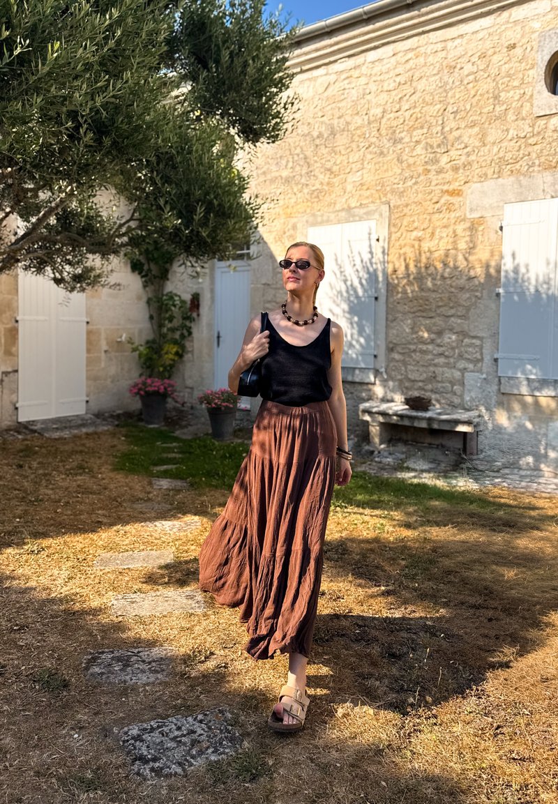 Donna che indossa un top nero, una lunga gonna marrone e sandali che cammina su un sentiero di pietra in un giardino soleggiato con muri di pietra e persiane bianche.