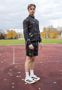 Sportkläder med en svart mönstrad jacka och matchande shorts, gjorda av lättviktigt tyg; kombinerade med vita skor med svarta ränder.