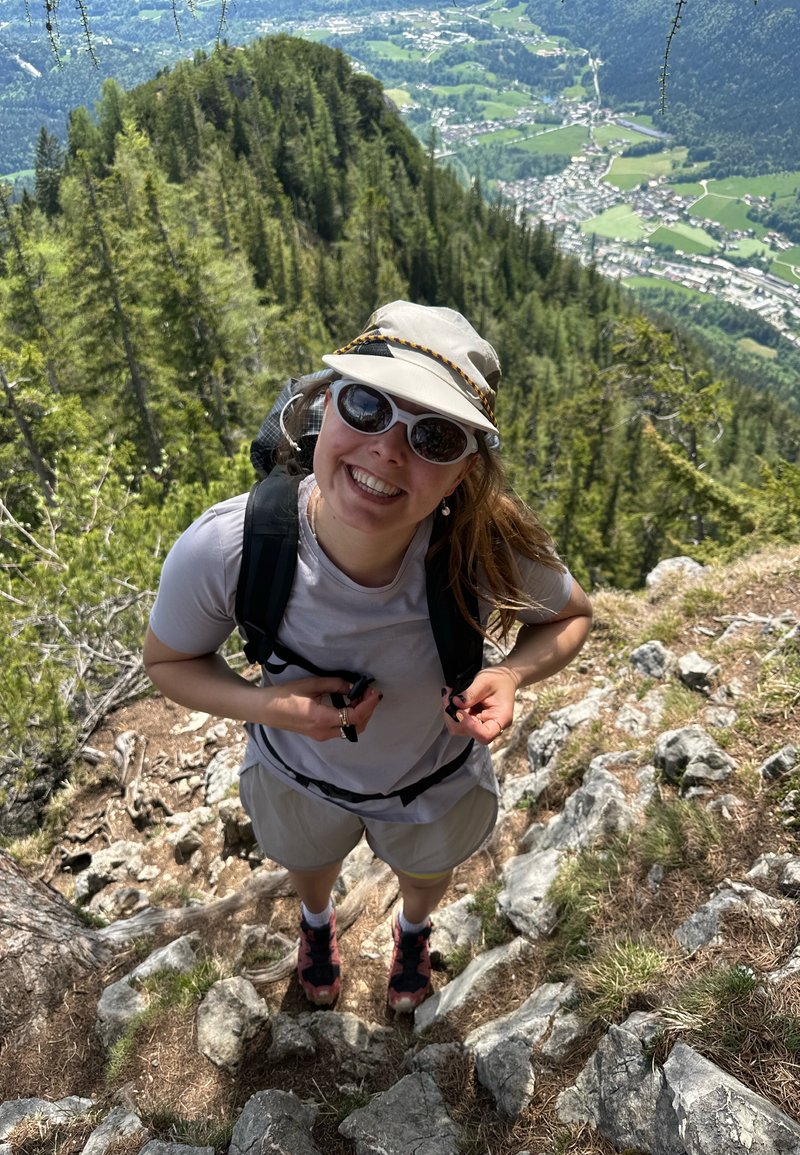 Femme avec lunettes de soleil et chapeau souriante en randonnée sur un sentier rocailleux avec forêt et village de la vallée en contrebas dans un cadre de montagne ensoleillé.