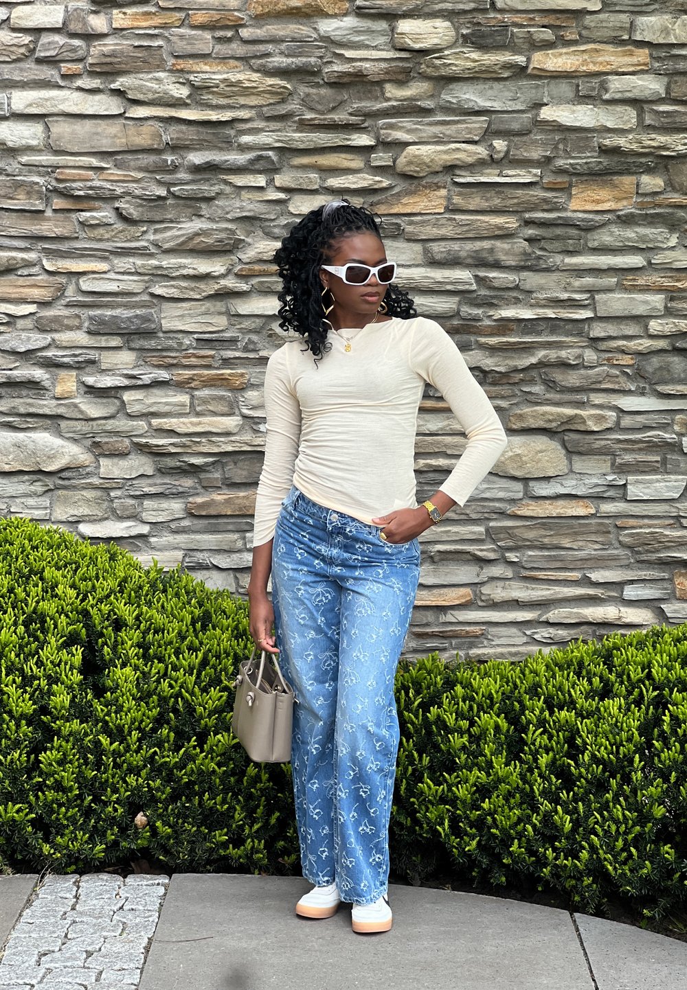 Blusa de mangas longas bege clara, jeans azuis com padrões florais brancos, óculos de sol brancos e uma bolsa cinza. De pé contra uma parede de pedras.