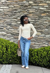 Blusa de mangas longas bege clara, jeans azuis com padrões florais brancos, óculos de sol brancos e uma bolsa cinza. De pé contra uma parede de pedras.