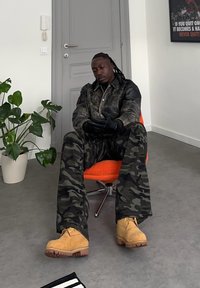 Homme aux cheveux tressés portant un pantalon camouflage et des bottes beige assis sur une chaise orange dans une pièce minimaliste avec une porte grise et une plante.