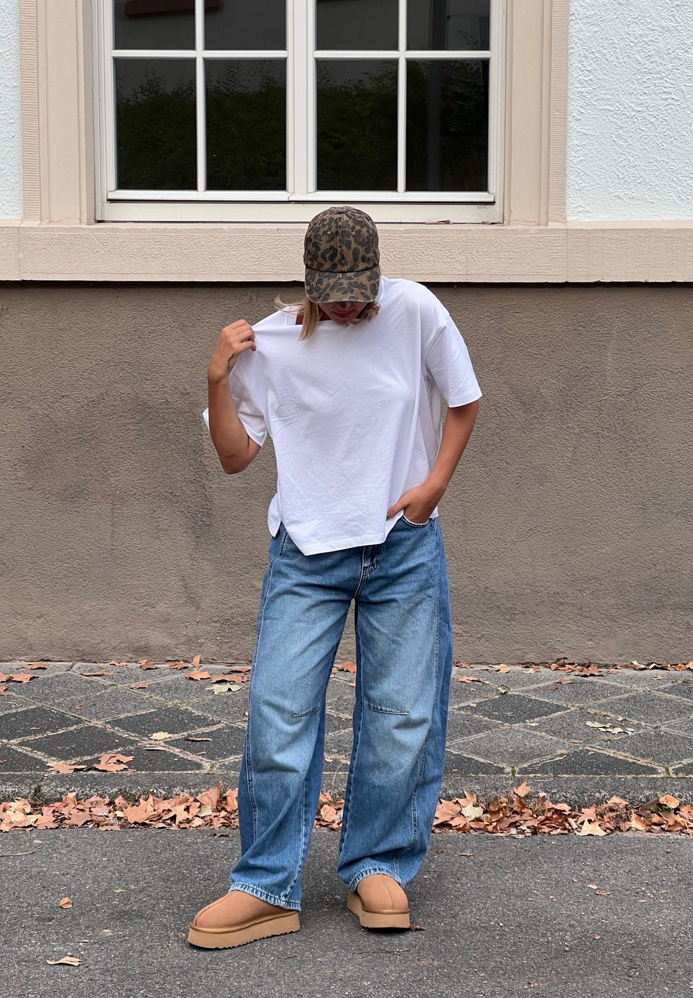 Maglietta bianca a maniche corte, vestibilità rilassata; jeans larghi azzurri; stivaletti beige slip-on; cappellino mimetico; ambiente esterno con foglie.