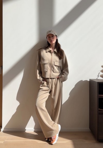 Mujer con chaqueta beige con cremallera y pantalones anchos con gorra beige y zapatillas deportivas, de pie contra una pared iluminada por el sol con sombras.