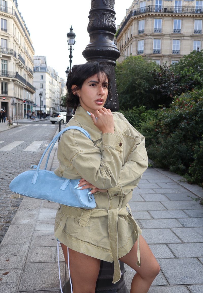 Femme aux cheveux courts et foncés appuyée contre un réverbère noir dans une rue de la ville, portant une veste beige et tenant un sac à main bleu clair.