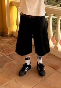 Svarta bomullsshorts, vit t-shirt, svarta sneakers med tre vita ränder, och vita ankelsockor med svarta ränder på en kaklad yta.