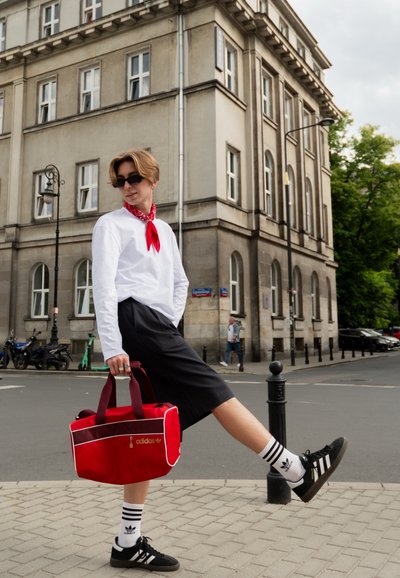 Bolsa de deporte roja de Adidas con asas color burdeos, forma rectangular, detalles de cremallera dorada, combinada con una camiseta de manga larga blanca y pantalones cortos negros.