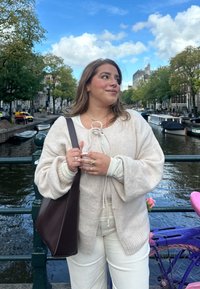 Beige oversized tröja med dragsnöre, lagd över en ljus topp, matchad med vita byxor. Brunt axelväska, ringar på fingrarna, kanal i bakgrunden.