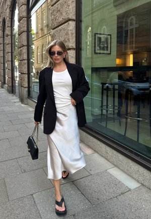 Blazer negro de corte oversized, top ajustado blanco, vestido de satén plateado tipo slip, bolso negro con cadena y chanclas negras gruesas.
