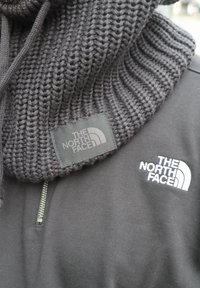 Czarny dziergany szalik o prążkowanej fakturze, z widoczną metką. Czarna bluza z kapturem z białym logo na piersi i suwakiem na ćwierć długości.