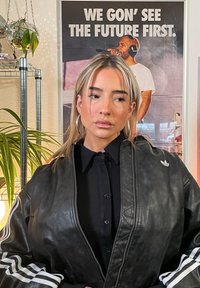 Casaco bomber de couro preto com punhos canelados, fecho de correr prateado e riscas brancas da Adidas. Usado sobre uma camisa preta de botões.